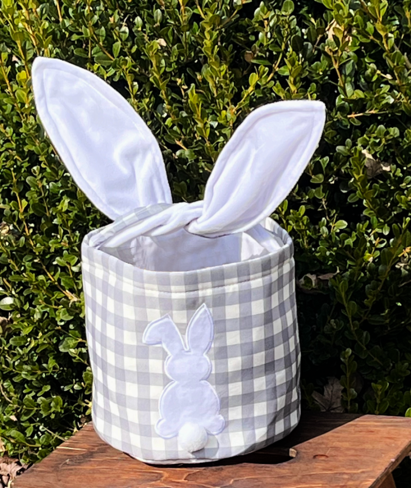 Grey checkered Easter Basket with hard wire ears for handle on brown bench with green tree background .Bunny on the front with Pom Pom tail sublimation Edit alt text