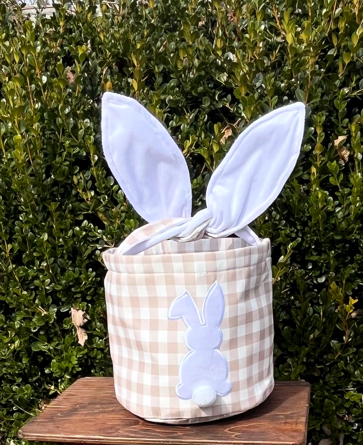 Tan checkered Easter Basket with hard wire ears for handle on brown bench with green tree background .Bunny on the front with Pom Pom tail sublimation