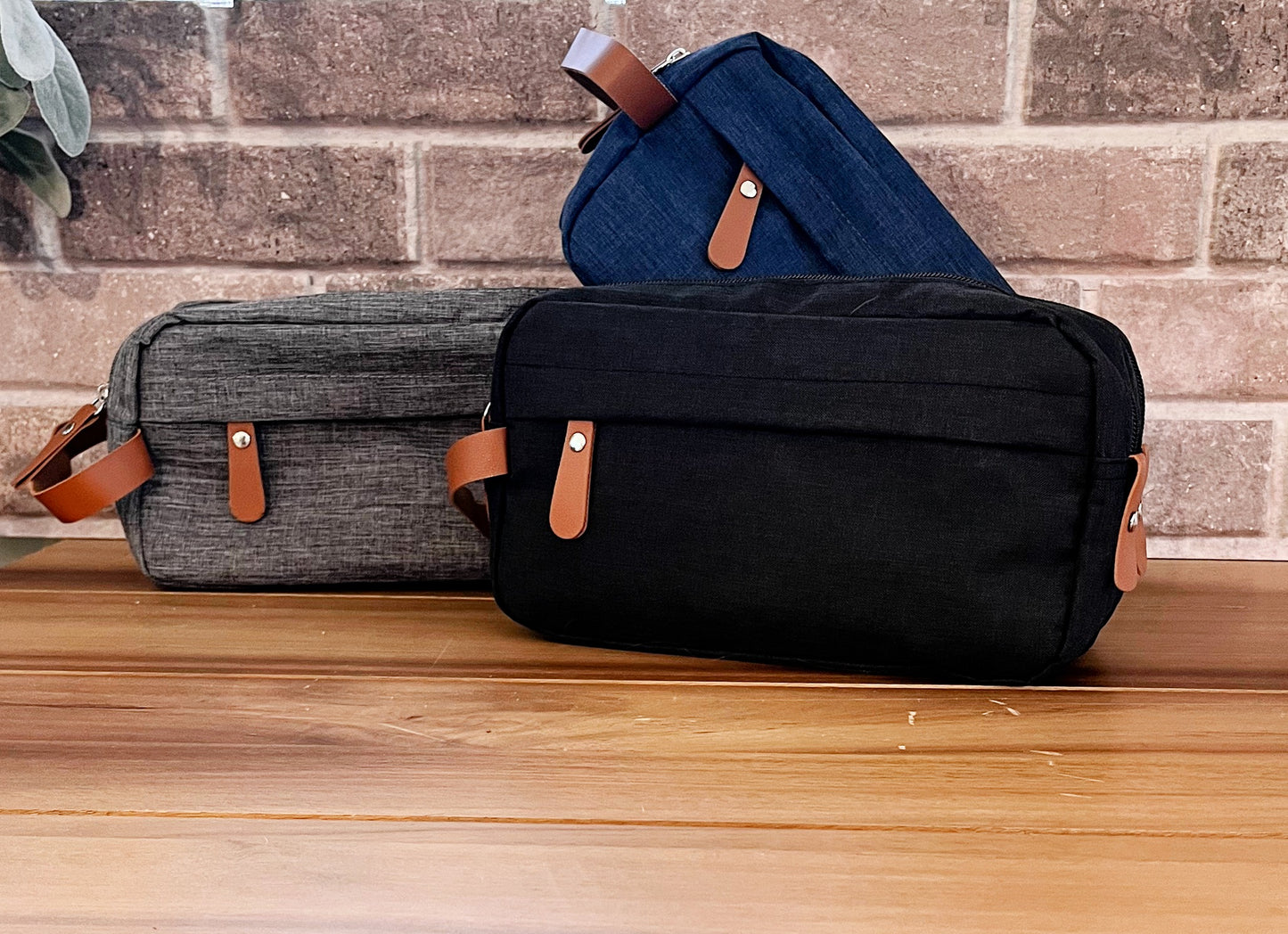 grey blue and black shaving bags with tan strap and zipper on wood table with brick background 