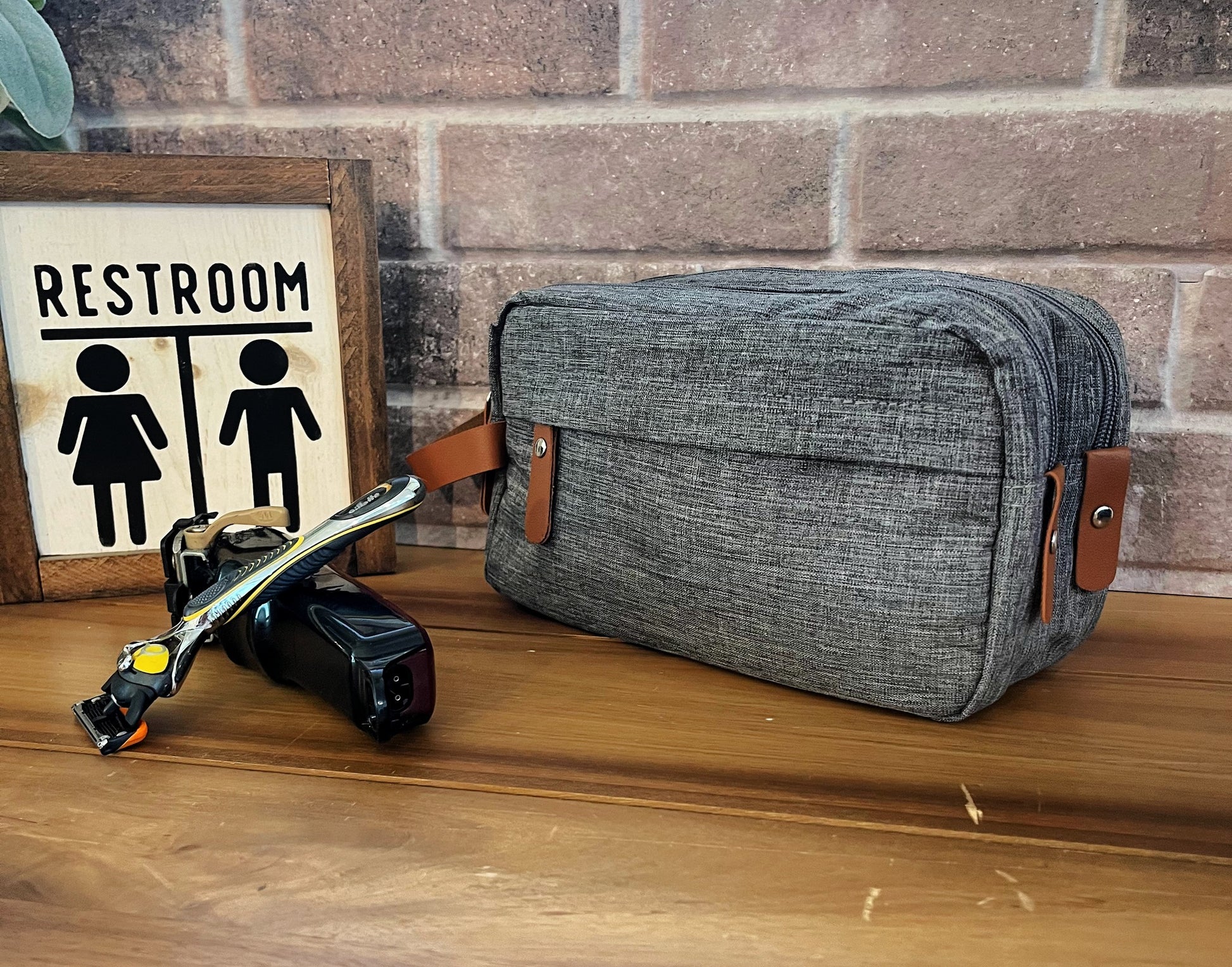 grey shaving bag with two zippers tan holder on wood table with razor bathroom sign and candle front of brick background