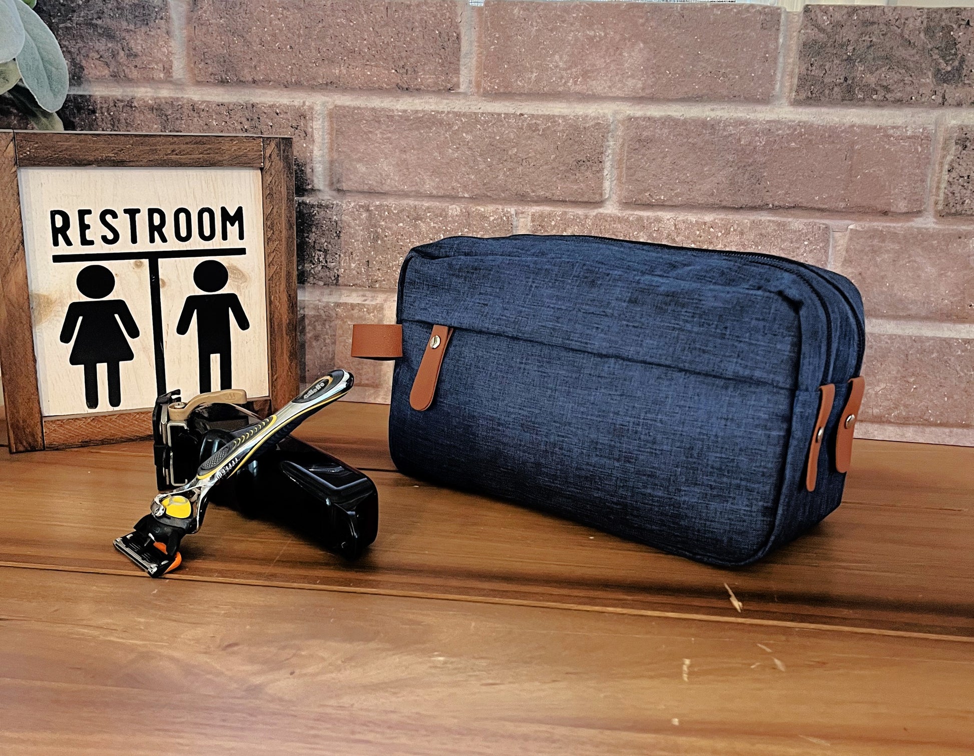 blue shaving bag with two zippers tan holder on wood table with razor bathroom sign in front of brick background