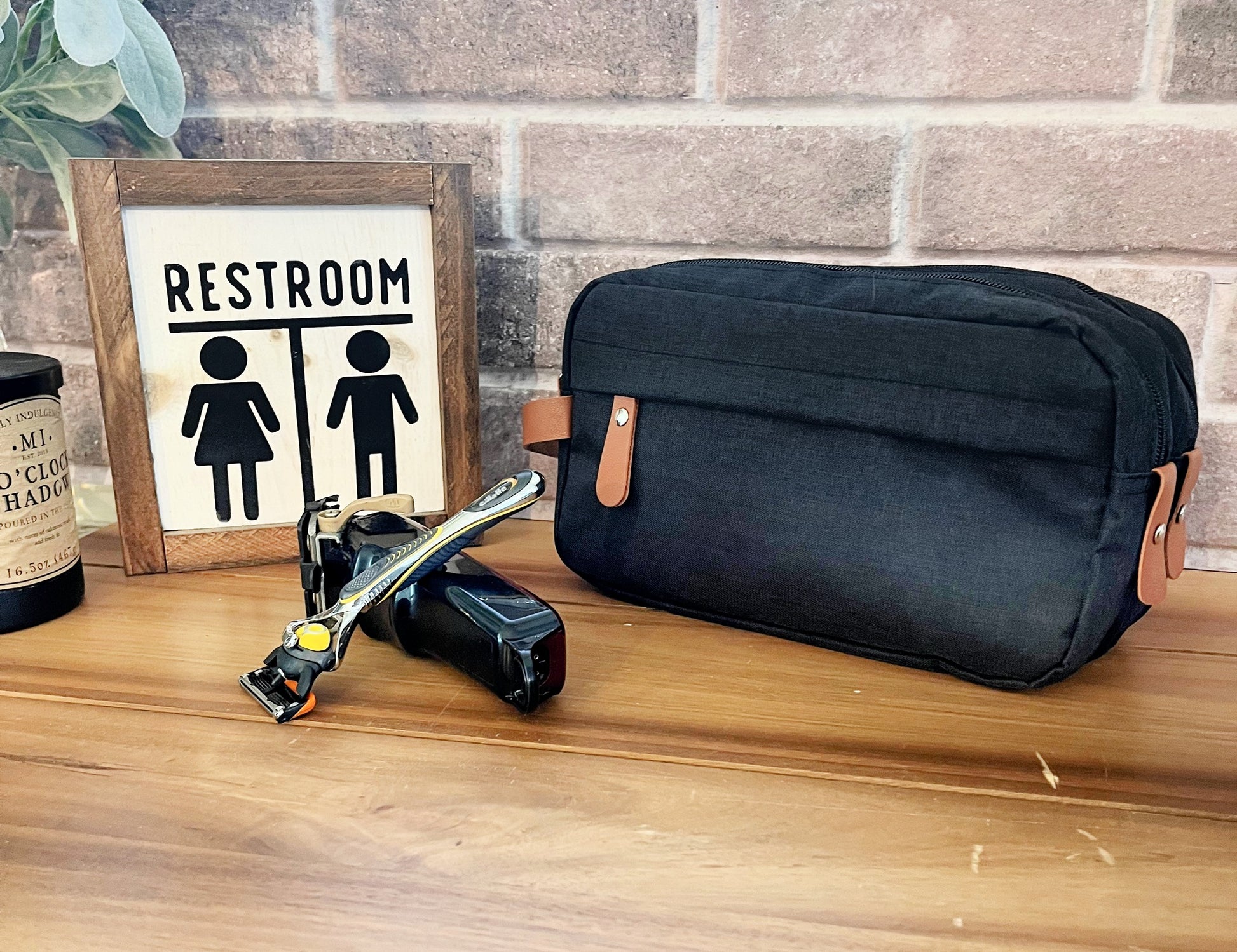 black shaving bag with two zippers tan holder on wood table with razor bathroom sign and candle front of brick background