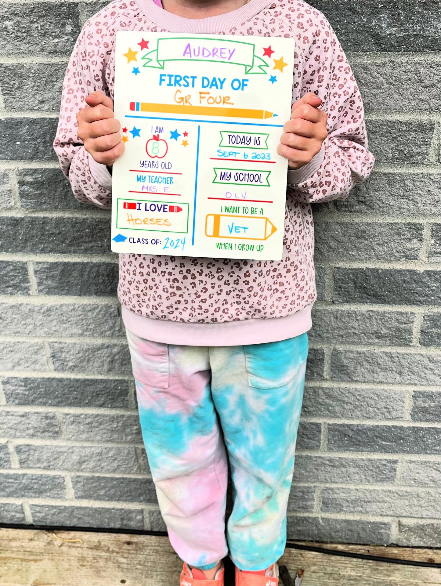 girl holding back to school personalized board with tie dye pants and cheetah sweater against brick background