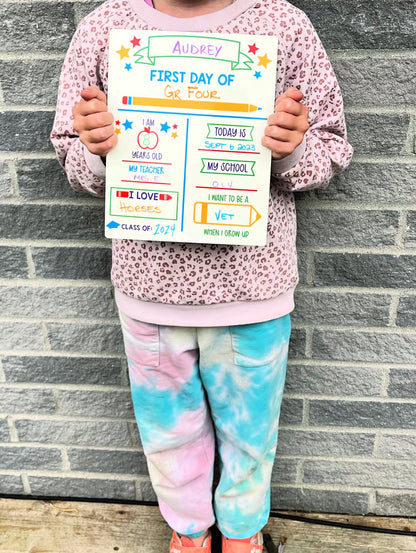 girl holding back to school personalized board with tie dye pants and cheetah sweater against brick background