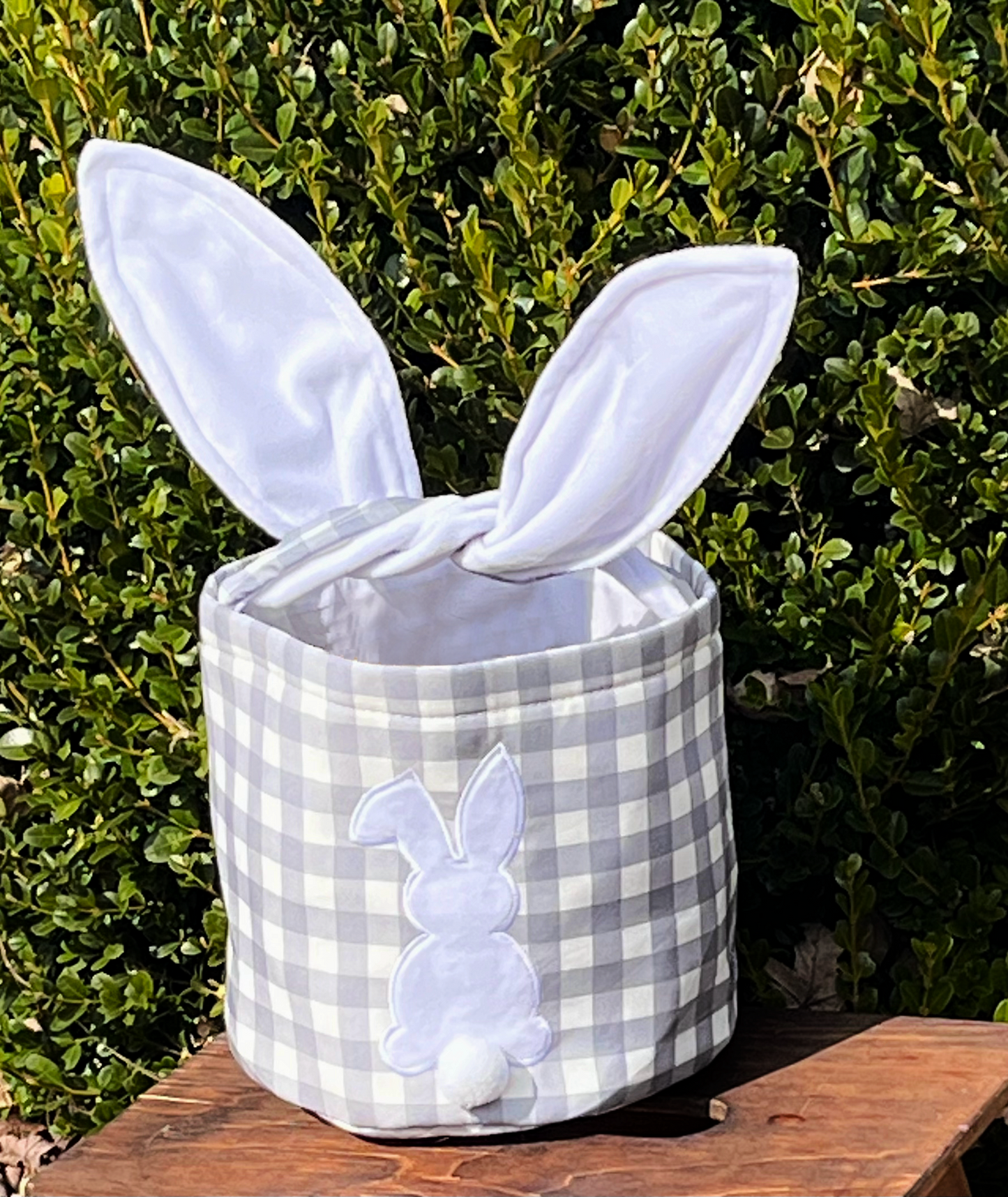 Grey checkered Easter Basket with hard wire ears for handle on brown bench with green tree background .Bunny on the front with Pom Pom tail sublimation 