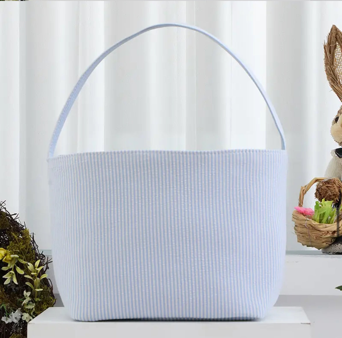 blue striped seer sucker Easter basket with handle on white stand with wreath and rabbit in background 