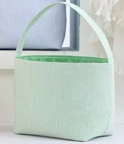 green striped seer sucker Easter basket with handle on white stand with blue basket  in background 