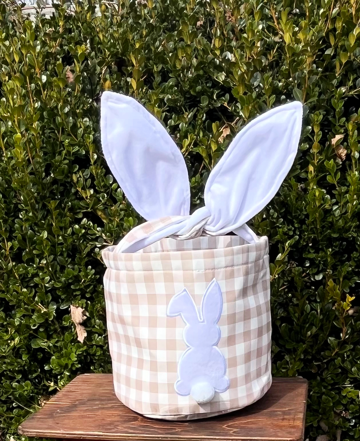 Tan checkered Easter Basket with hard wire ears for handle on brown bench with green tree background .Bunny on the front with Pom Pom tail sublimation 