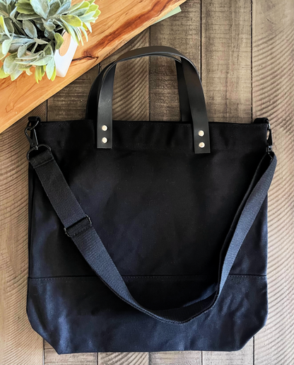 Leather handle black canvas bag with black strap on wood background with green leaves