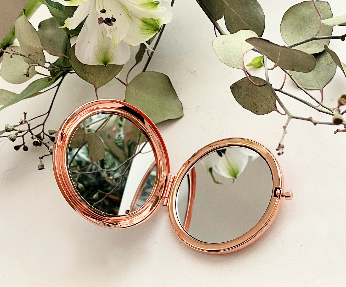 rose gold compact mirror open on white table with green leaves and white flowers